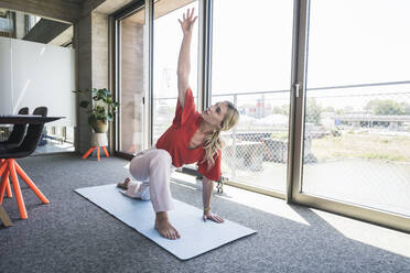 Businesswoman with hand raised practicing yoga on exercise mat in office - UUF26899