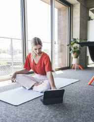 Businesswoman using tablet PC sitting on exercise mat in office - UUF26900