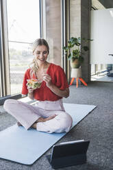 Smiling businesswoman eating salad watching tablet PC on exercise mat in office - UUF26901