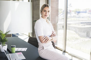 Young businesswoman with arms crossed sitting on desk in office - UUF26913