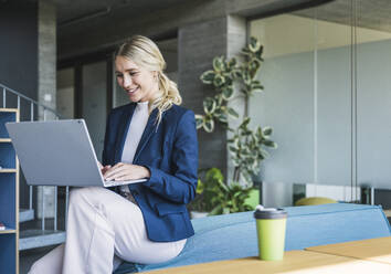 Smiling businesswoman using laptop sitting on couch in office - UUF26923