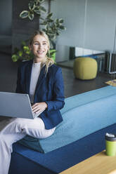 Smiling businesswoman sitting with laptop on couch in office - UUF26925