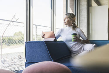 Businesswoman with tablet PC and reusable cup leaning on couch in office - UUF26929