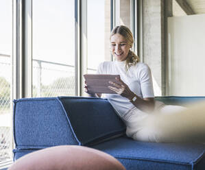 Happy businesswoman using tablet PC leaning on couch in office - UUF26931