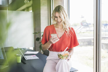 Businesswoman using smart phone holding lunch box sitting on desk in office - UUF26962