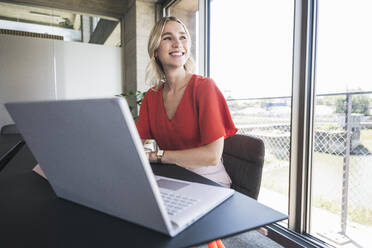Glückliche blonde Geschäftsfrau sitzt mit Laptop am Schreibtisch im Büro - UUF26976
