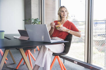 Junge Geschäftsfrau mit Lunchpaket auf einem Stuhl im Büro sitzend - UUF26979