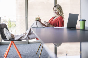 Lächelnde Geschäftsfrau mit Lunchbox und Smartphone auf einem Stuhl im Büro sitzend - UUF26984