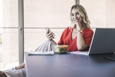 Happy businesswoman with hand on chin holding smart phone sitting at desk in office - UUF26987