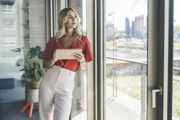 Young businesswoman with tablet PC leaning on window in office - UUF26995