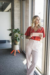 Businesswoman with tablet PC leaning on window in office - UUF26996