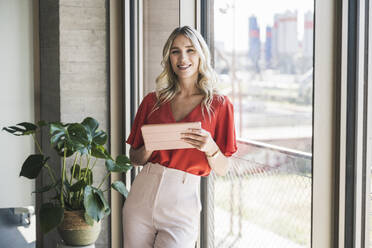 Lächelnde blonde Geschäftsfrau mit Tablet-PC, die sich an ein Fenster im Büro lehnt - UUF26998