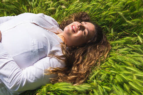 Von oben optimistische Frau auf grünem Gras liegend mit geschlossenen Augen beim Ausruhen an einem sonnigen Sommertag in der Natur - ADSF35926