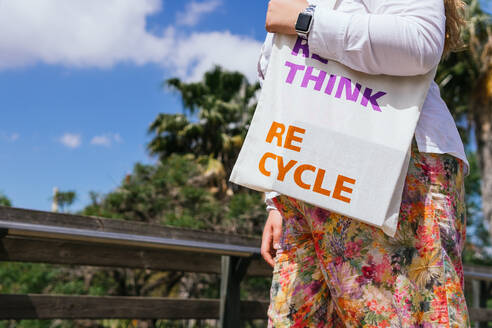 Crop anonymous female carrying bag with Re Think Re Cycle inscription while standing near fence on street on summer day - ADSF35927
