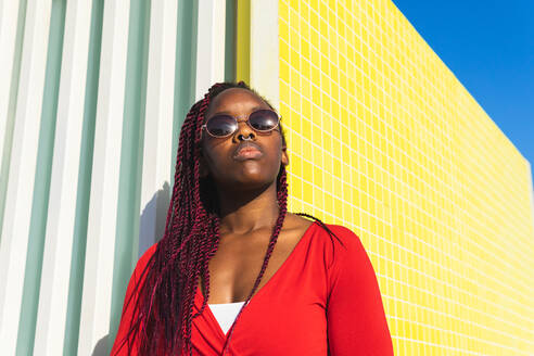 From bellow young African American female in trendy outfit and glasses looking away while leaning against colorful wall on a sunny day - ADSF35934