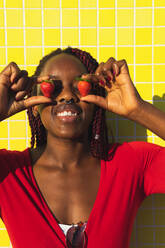 Young African American female in trendy red outfit looking at camera while holding strawberries in front of face - ADSF35937