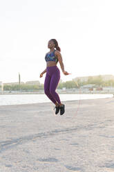 Full body fit black woman athlete in sportswear jumping with rope at the sea shore - ADSF35946