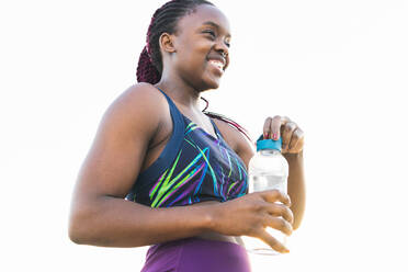 Seitenansicht einer afroamerikanischen Sportlerin mit einer Wasserflasche, die lächelt und wegschaut, während sie auf einem weißen Hintergrund steht - ADSF35950
