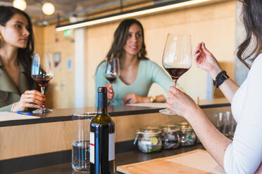 Weibliche Sommeliers in eleganter Freizeitkleidung genießen das Aroma von Rotwein, während sie an der Theke einer Bar mit einem nicht erkennbaren Barkeeper sitzen - ADSF35956