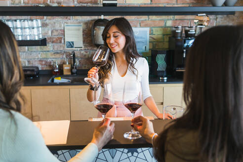 Weibliche Barkeeperin in eleganter Freizeitkleidung genießt das Aroma von Rotwein, während sie während einer Degustation in einer Bar mit unkenntlichen weiblichen Sommeliers an der Theke sitzt - ADSF35957