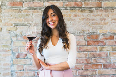 Ethnische Frau in trendigem Outfit mit einem Glas Rotwein, lächelnd und in die Kamera blickend, während sie in einem Restaurant an einer Backsteinmauer steht - ADSF35961