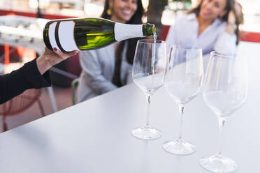 Abgeschnittene, nicht erkennbare Person, die Weingläser mit weißem Sekt füllt, während sie mit Freundinnen an einem Tisch auf einer Restaurantterrasse sitzt - ADSF35964