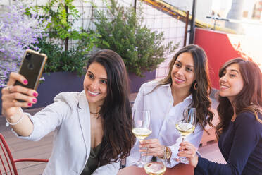 From above of adult Hispanic women with glasses of white wine smiling and taking selfie via smartphone while spending time on restaurant terrace on weekend day - ADSF35971