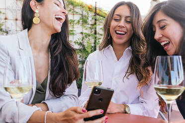 Erwachsene hispanische Frauen mit einem Glas Weißwein, die lächelnd auf ihrem Smartphone surfen, während sie am Wochenende auf der Terrasse eines Restaurants Zeit verbringen - ADSF35973