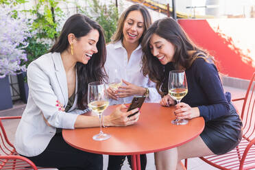 Adult Hispanic women with glasses of white wine smiling and browsing on smartphone while spending time on restaurant terrace on weekend day - ADSF35974