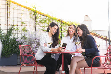 Adult Hispanic women with glasses of white wine smiling and browsing on smartphone while spending time on restaurant terrace on weekend day - ADSF35975