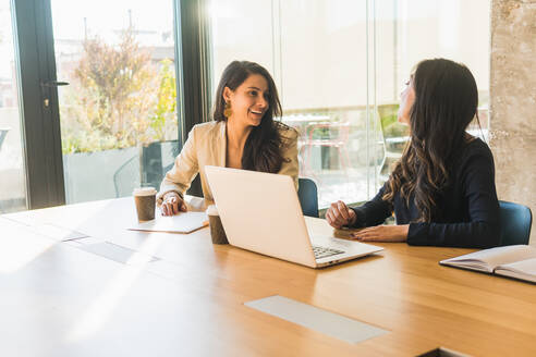 High angle of cheerful Hispanic female entrepreneurs in smart casual clothes smiling and discussing business ideas while sitting at table and working on project in sunlit office together - ADSF35983