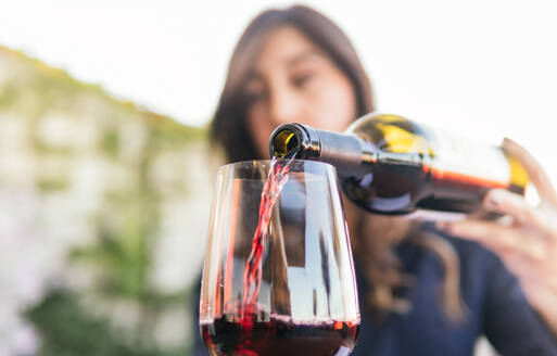 Cropped unrecognizable ethnic woman with long hair filling goblet with red wine from bottle with blank label during degustation session in restaurant - ADSF35986