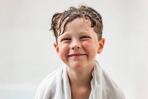 Lächelndes Kind, das an der Badewanne steht und in die Kamera schaut, nachdem es in einem weißen Badezimmer zu Hause Hygienemaßnahmen durchgeführt hat - ADSF36015