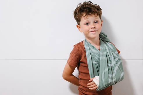 Positive cute boy with broken arm in cloth sling holding hand behind back and looking at camera with smile while standing against white wall - ADSF36016