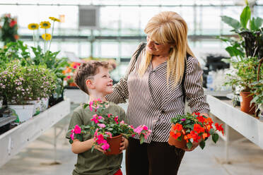 Glückliche blonde Oma mit Impatiens Pflanze lächelnd und Blick auf Enkelkind beim Besuch Blumenladen am Wochenende Tag zusammen - ADSF36024