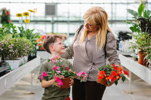 Happy blond grandma with impatiens plant smiling and looking at grandchild while visiting flower shop on weekend day together - ADSF36024