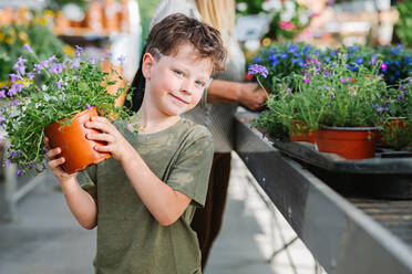 Niedliches Kind, das eine Topfpflanze mit lila Blume trägt und in die Kamera schaut, während es in der Nähe eines Verkaufsstandes steht - ADSF36025