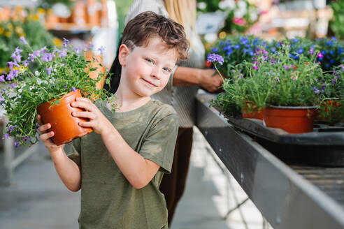 Cute child carrying potted plant with purple flower and looking at camera while standing near stall in shop - ADSF36025