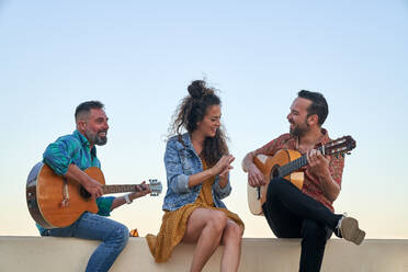 Zufriedene männliche Musiker spielen ein Lied auf akustischen Gitarren in der Nähe einer klatschenden Frau, die am Rande einer Stadt vor einem blauen wolkenlosen Himmel sitzt - ADSF36046