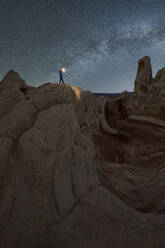 Silhouette eines nicht erkennbaren Forschers, der mit einer Taschenlampe auf einer Landschaft von Felsformationen im Hochland unter einem milchigen Sternenhimmel im Vermillion Cliffs National Monument, Arizona in den USA steht - ADSF36058