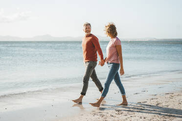 Happy mature couple holding hands walking on shore at beach - JOSEF11291