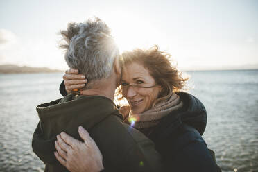 Smiling mature woman embracing man at beach on sunny day - JOSEF11296