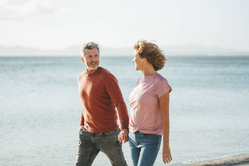 Glücklicher Mann und Frau halten sich an den Händen und gehen am Strand spazieren - JOSEF11308