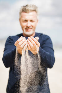 Lächelnder Mann mit Sand in den Händen am Strand - JOSEF11315