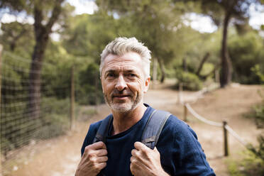 Smiling mature man with backpack standing in front of trees - JOSEF11334