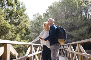 Mature couple with smart phone standing on footbridge - JOSEF11336
