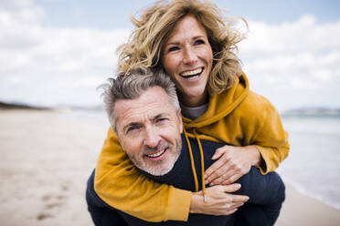 Happy man giving piggyback ride to woman at beach - JOSEF11357