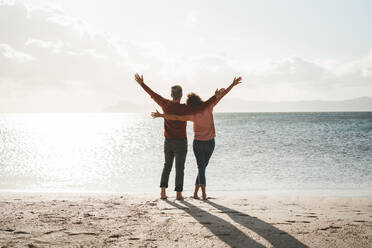 Carefree couple with arms outstretched standing on shore at beach - JOSEF11390