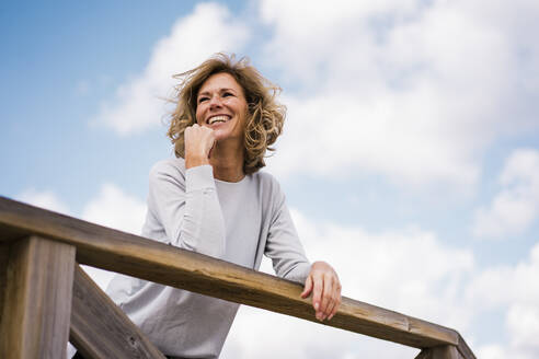 Happy mature woman leaning on railing under cloudy sky - JOSEF11396