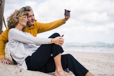 Lächelnder Mann nimmt Selfie mit Frau durch Handy am Strand - JOSEF11402